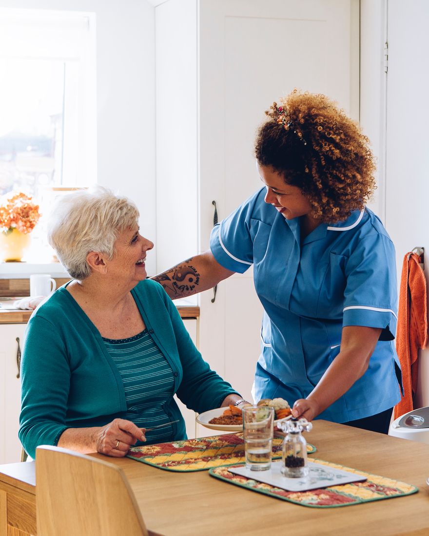 A smiling carer serving lunch to an older lady at her kitchen table
